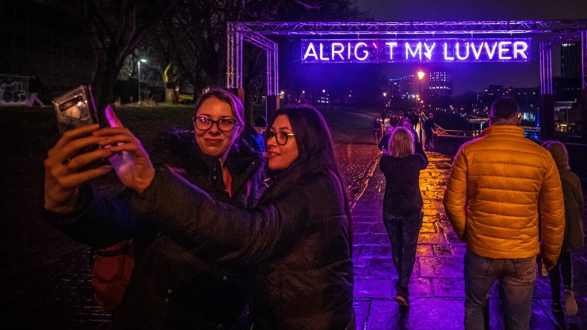 Two people taking a selfie in front of a neon sign that read: Alright my luvver, at Bristol Light Festival