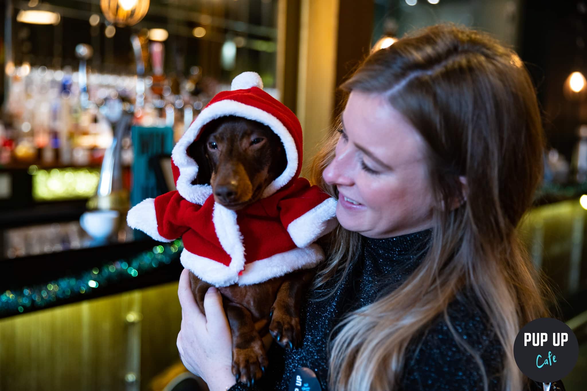 A woman holding a dachshund that is dressed as Father Christmas.