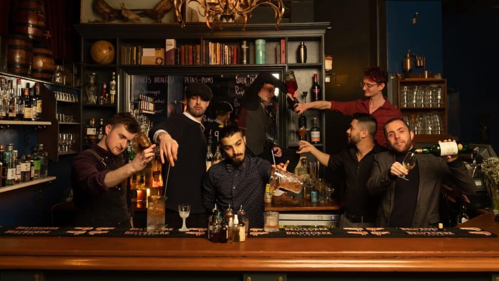 Six of The Milk Thistle's bartenders making cocktails around the bar, surrounded by books, taxidermy birds and other antiques