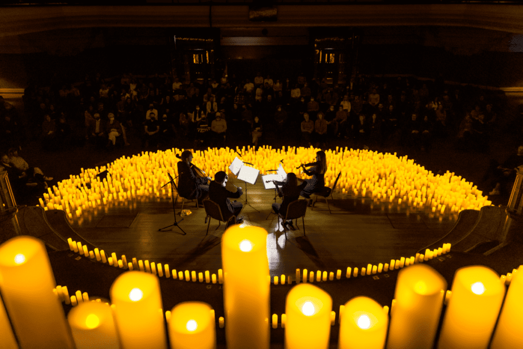 A string quartet performing on a stage surrounded by candles with the audience visible in the background and a close-up of seven candles in the foreground.