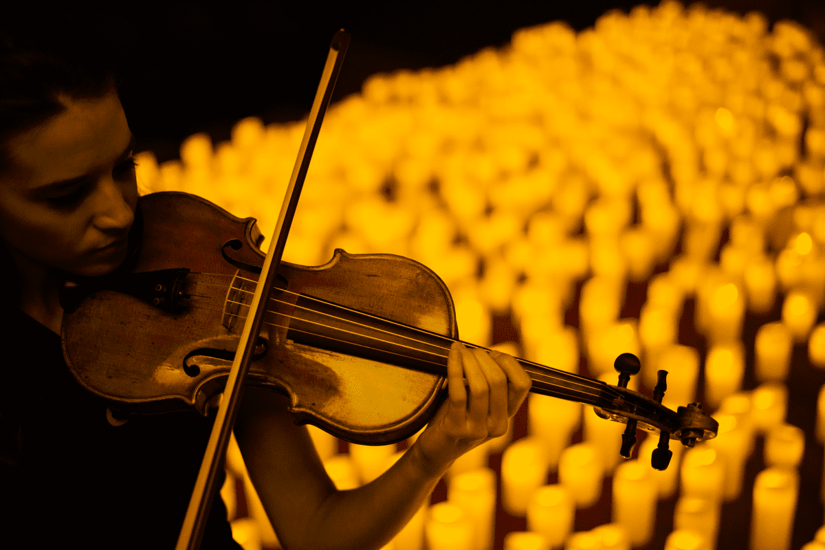 A close-up of a woman playing the violin with a sea of candles behind her.