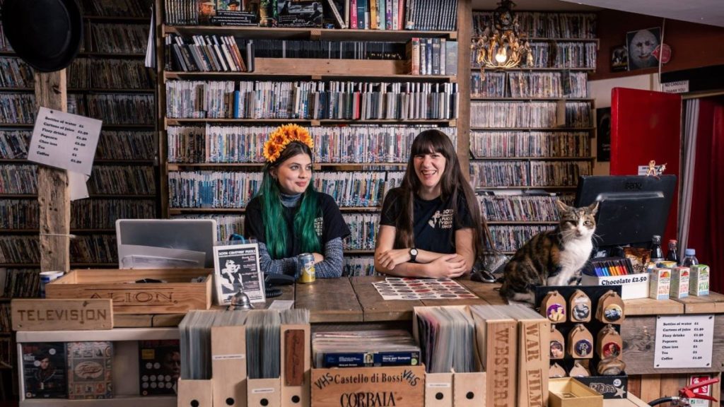 inside of 20th century FLicks, with two staff at the counter and a cat, surrounded by shelves of DVDs